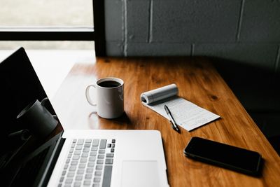 Workspace with laptop, coffee, notepad, pen, and smartphone on wooden desk.