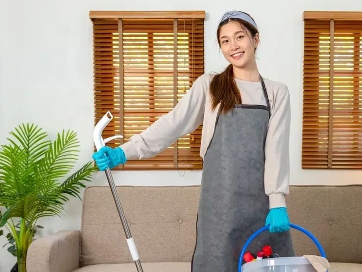 Smiling woman in apron holding cleaning supplies and mop in a tidy living room.