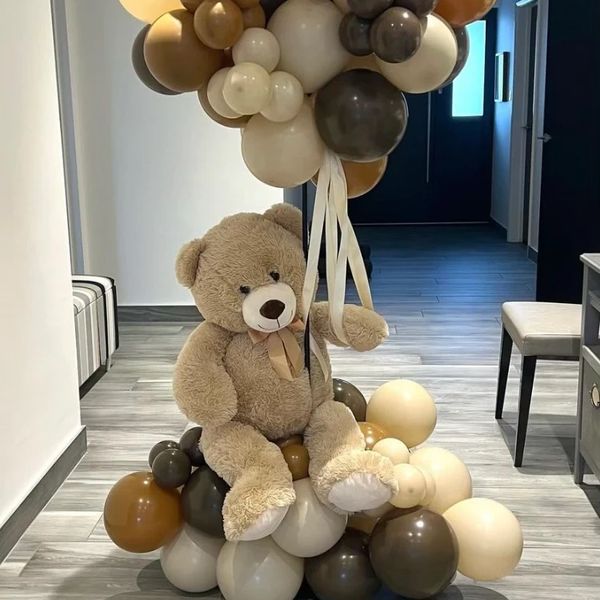 Large teddy bear holding brown and beige balloons indoors.