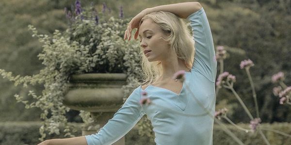 A woman in a light blue dress gracefully dances outdoors near flowers and greenery.