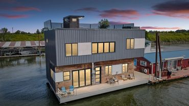 Modern floating house with large windows and outdoor seating on calm water.