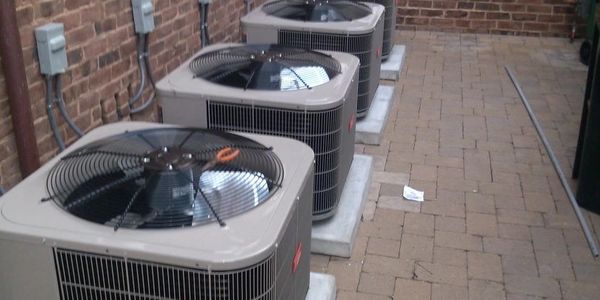 Three outdoor air conditioning units lined up beside a brick wall.