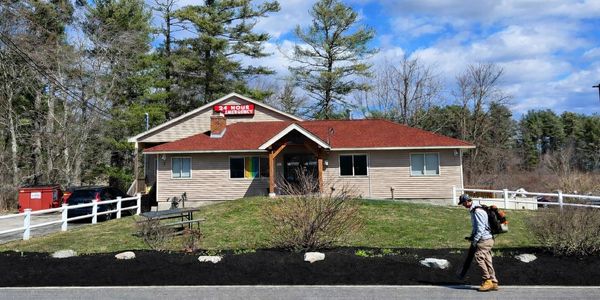 A person using a leaf blower in front of a building with a red roof on a sunny day.