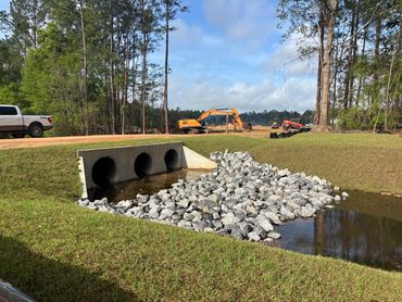 Concrete culvert with rocks by a grassy field and machinery in the background.