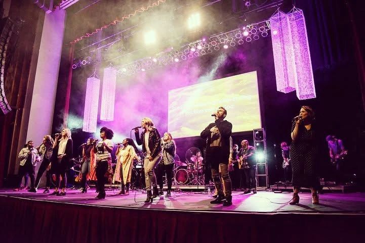 A large group of singers and musicians perform on a brightly lit stage with purple lighting.