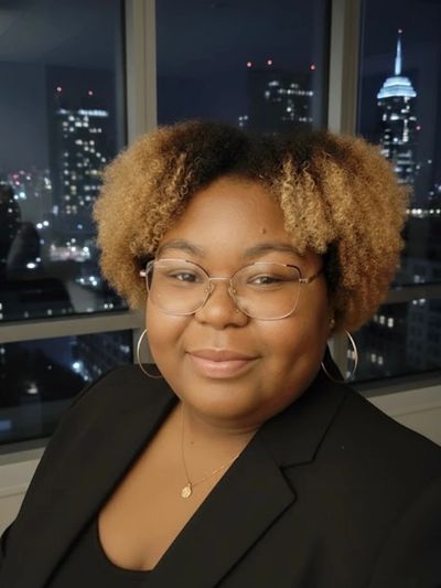 A woman with curly hair and glasses smiling indoors with a city skyline at night in the background.