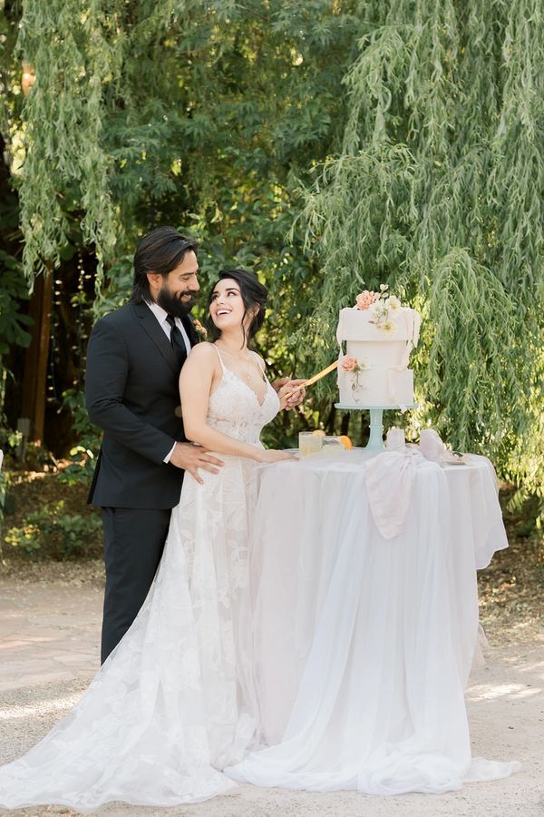 Bride and groom joyfully cut their wedding cake outdoors under lush greenery.