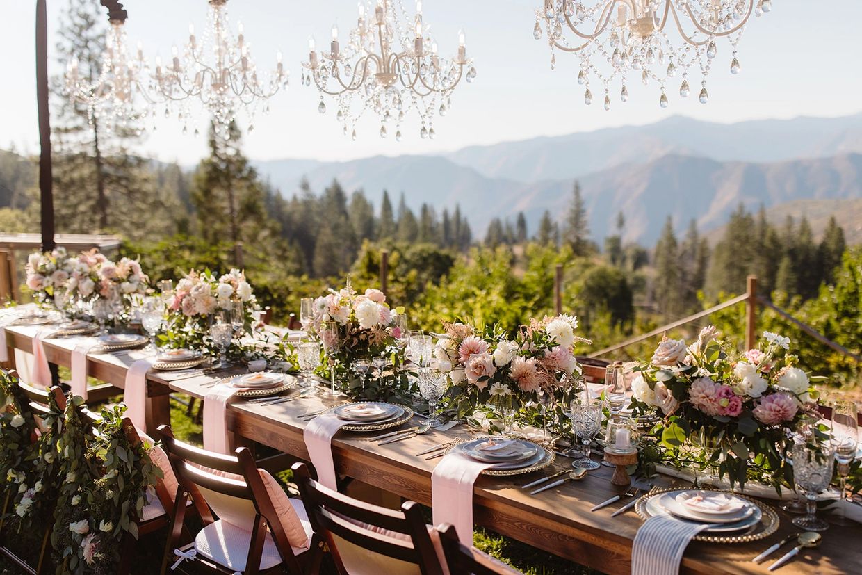 Elegant outdoor dining setup with floral arrangements and chandeliers against a mountain backdrop.