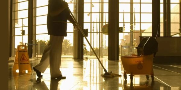 A janitor mops a shiny floor in a large, sunlit building.