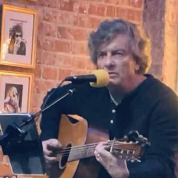 Man playing acoustic guitar and singing into a microphone indoors.