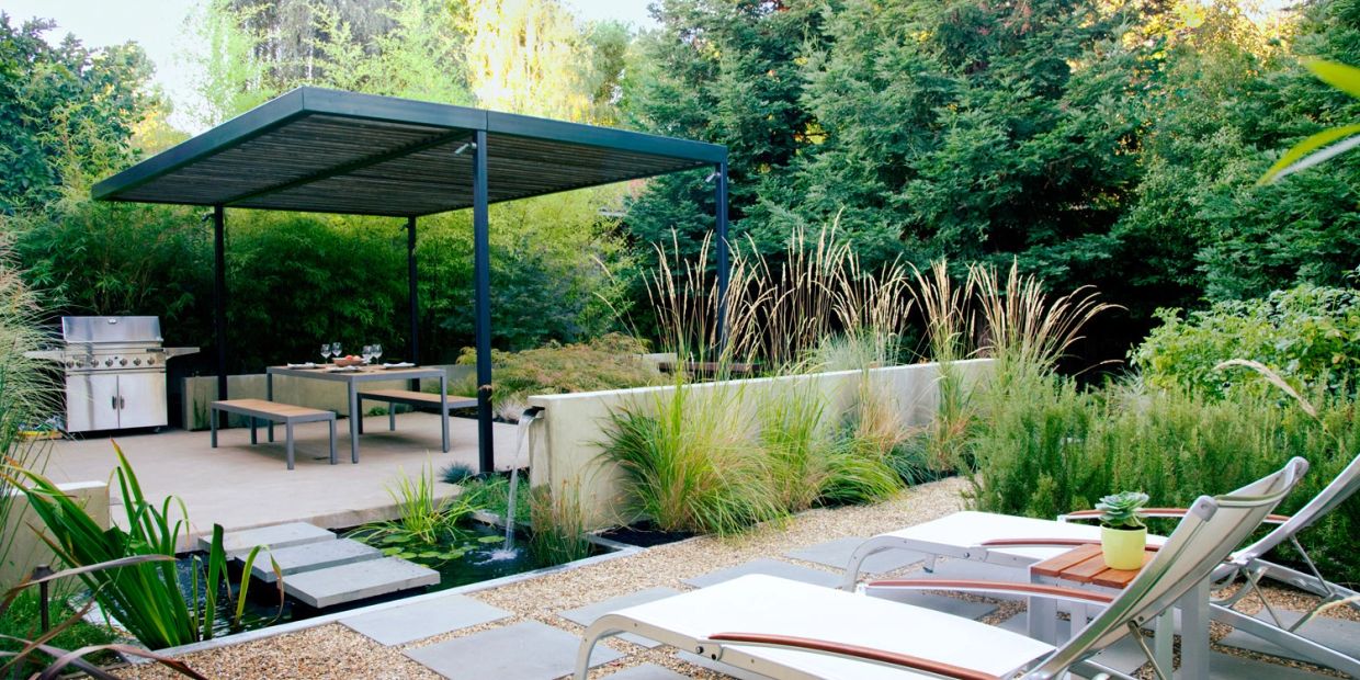 Modern outdoor patio with dining area, grill, water feature, and lounge chairs surrounded by lush greenery.