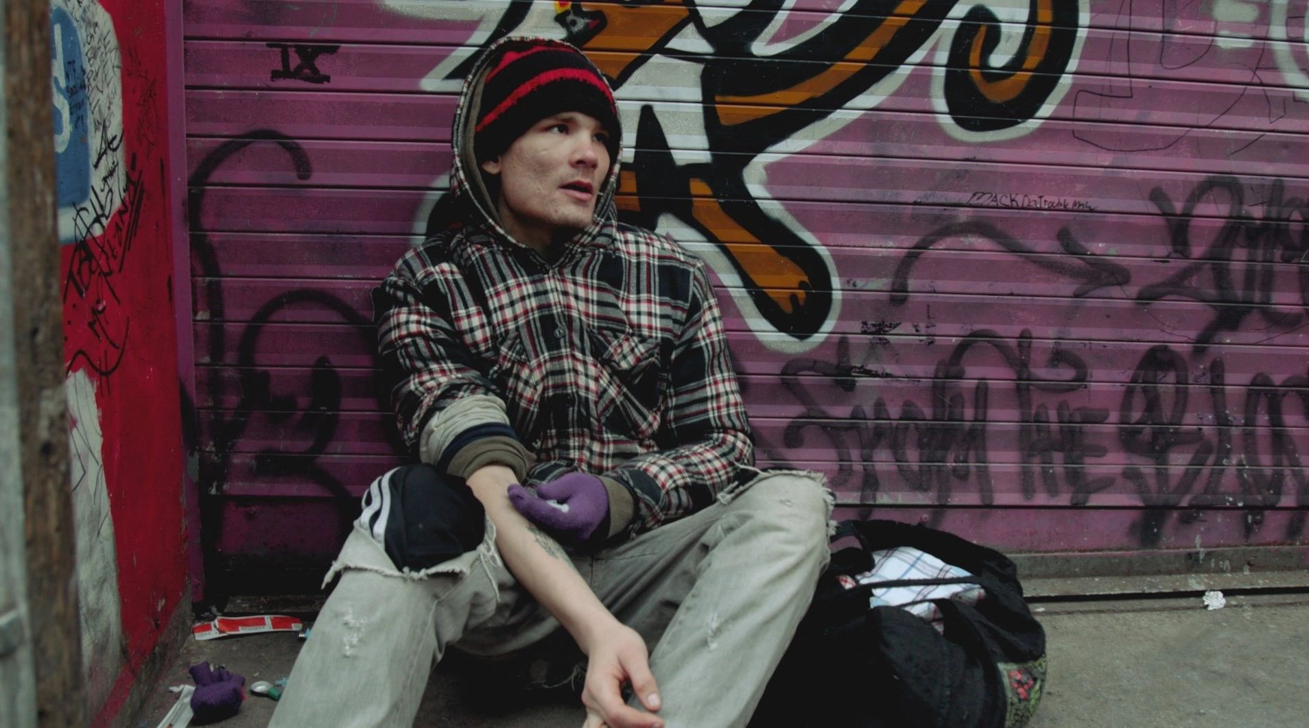 A young man in a beanie and plaid shirt sits against a graffiti-covered wall.