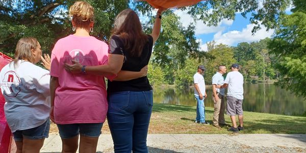 Three women standing arm in arm near a lake, with three men talking in the background.