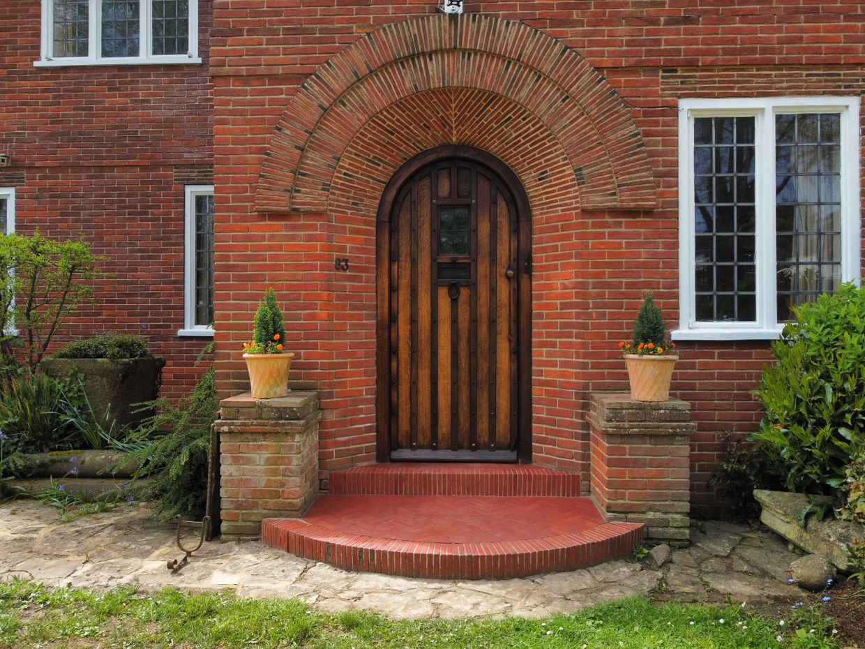 Arched wooden door framed by red bricks and two potted plants at the entrance.