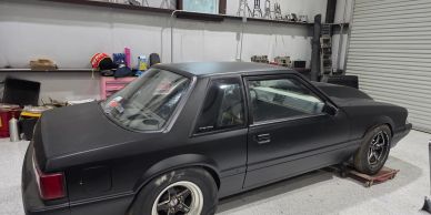 Rear-three-quarter view of a customized matte black Foxbody Ford Mustang parked inside a garage work