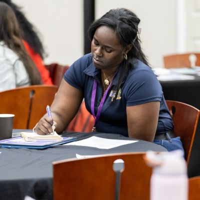 A woman attentively writes notes at a table during a meeting or workshop.