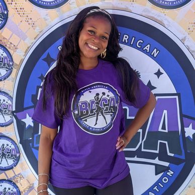 A smiling woman in a purple BACA shirt poses confidently in front of a BACA backdrop.