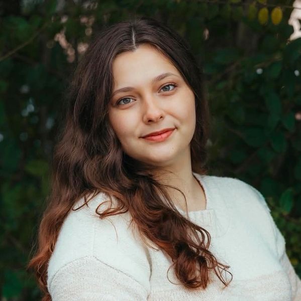Woman with long brown hair in white sweater smiling outdoors.