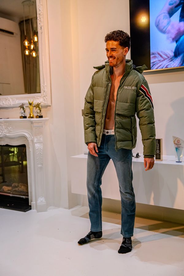 Man wearing a green jacket, jeans, and socks, smiling indoors.