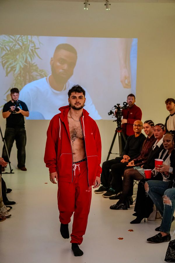 Male model in red tracksuit walking down runway during fashion show with audience and cameras.