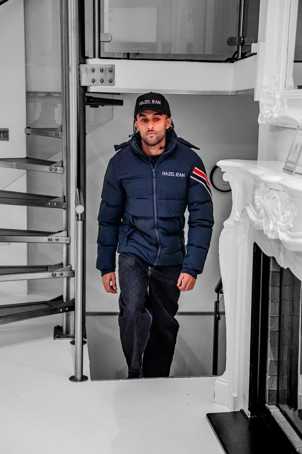 Man wearing a navy blue jacket and cap with "HAZEL JEAN" walking indoors.