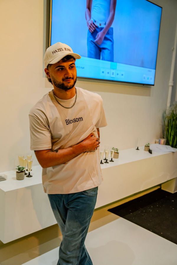 Man wearing a 'Blossom' shirt and 'HAZEL JEAN' cap stands indoors with a TV displaying a person in jeans behind him.