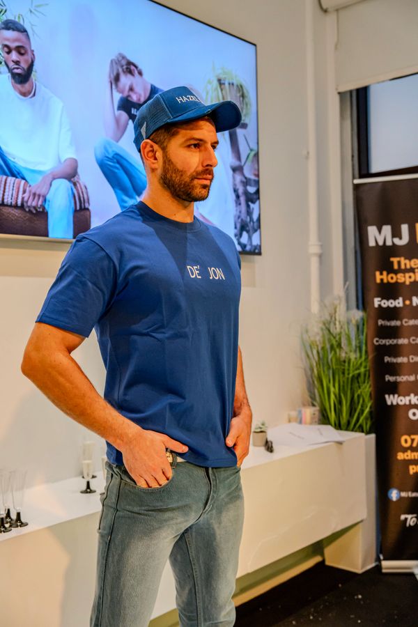A man in a blue cap and t-shirt poses indoors with hands in pockets.
