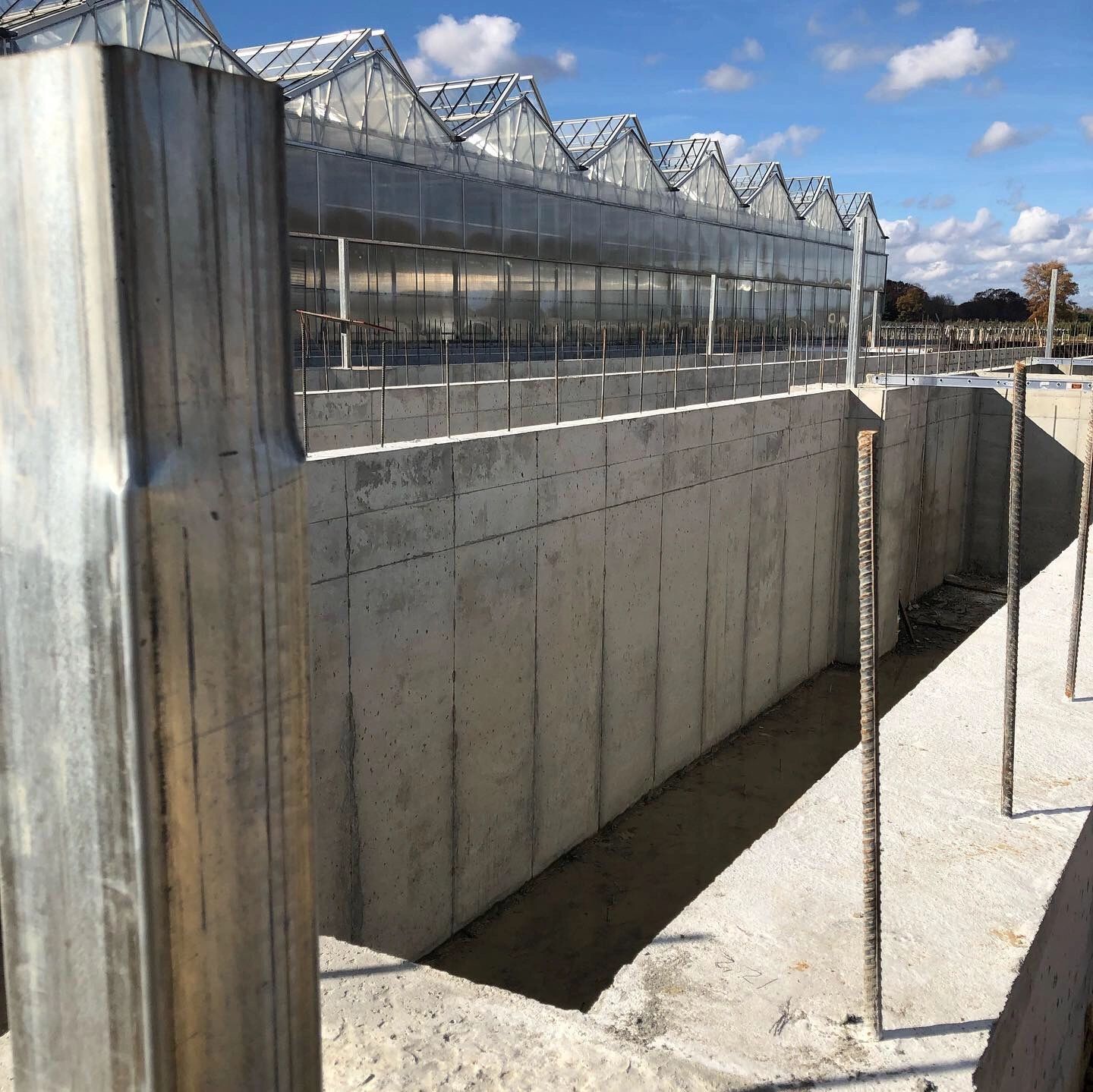 Construction site with concrete foundation and greenhouses in the background.