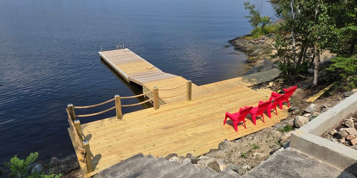 Wooden dock with red chairs by calm water surrounded by trees.