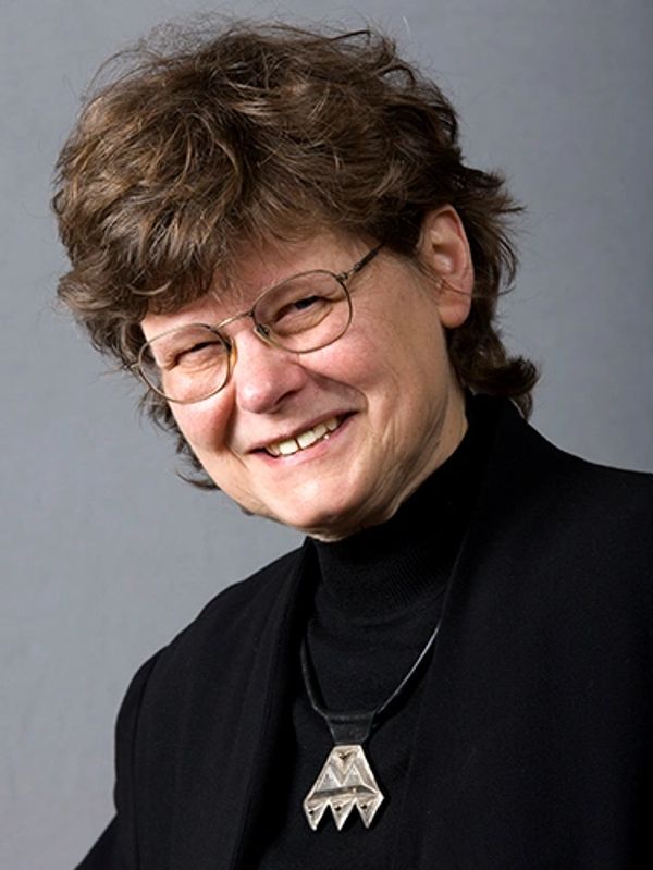 Smiling person with glasses wearing a black outfit and geometric pendant.