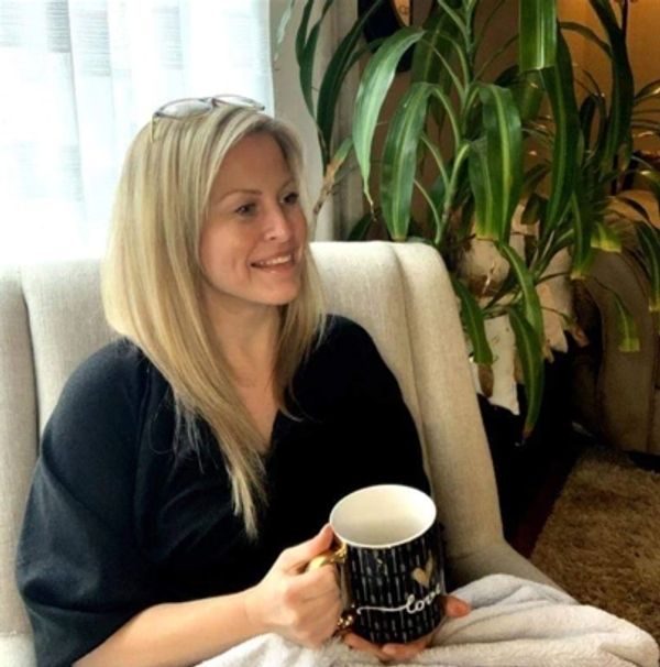 Woman with blonde hair smiling while holding a cup, sitting on a chair indoors.