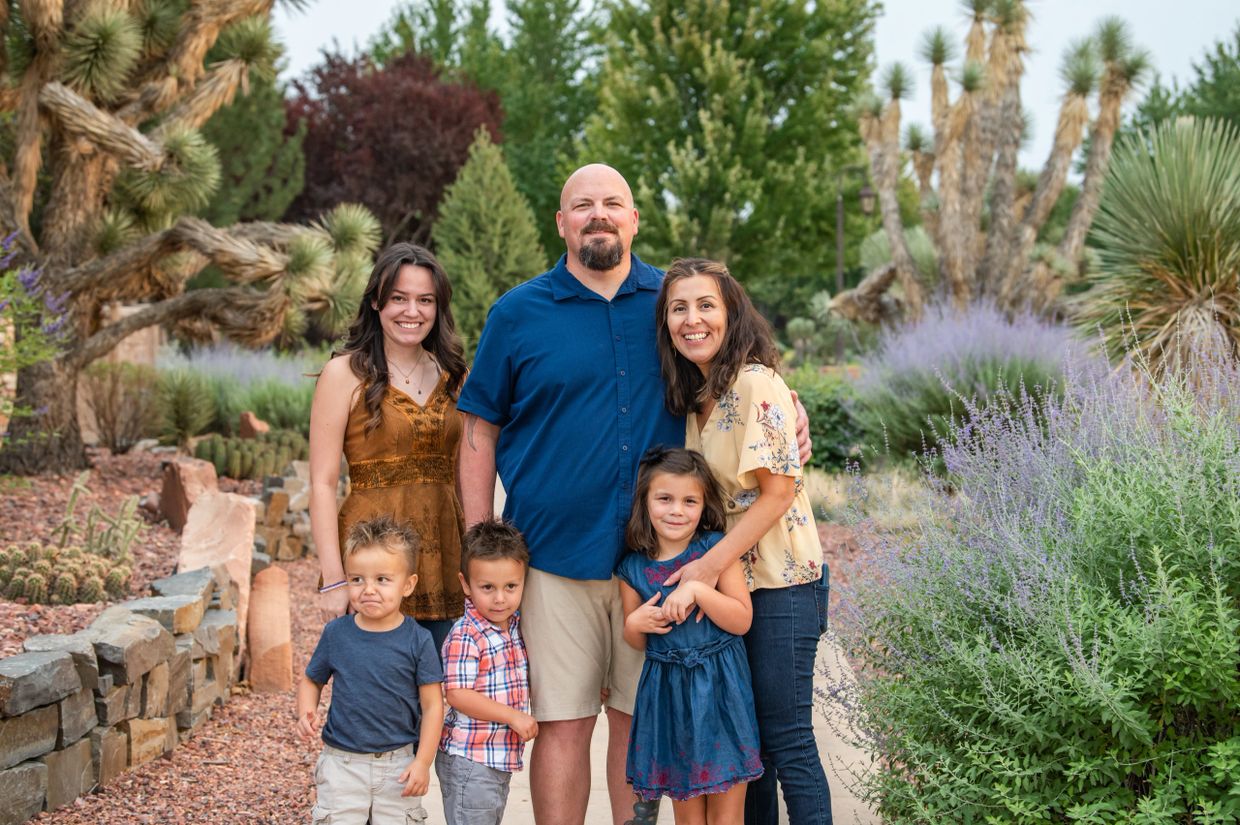 A happy family of six posing outdoors in a garden with greenery and flowers.