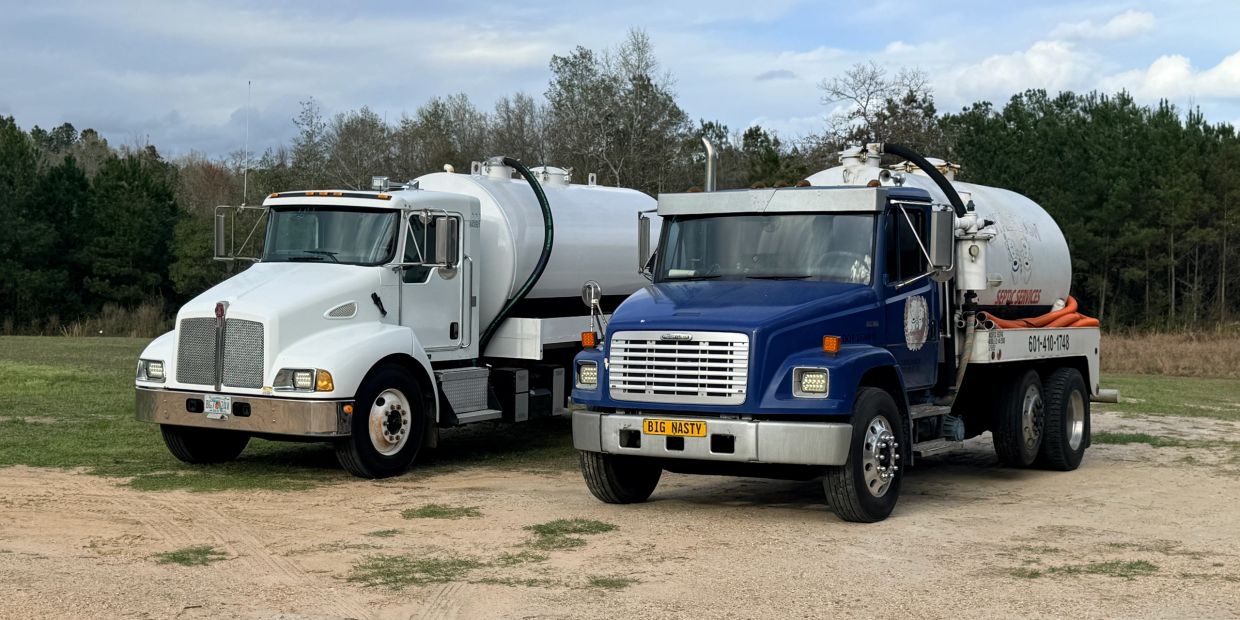 Two tanker trucks parked on dirt with trees behind.