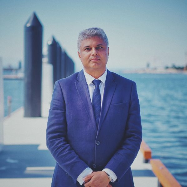 Man in blue suit standing near waterfront with a calm expression.
