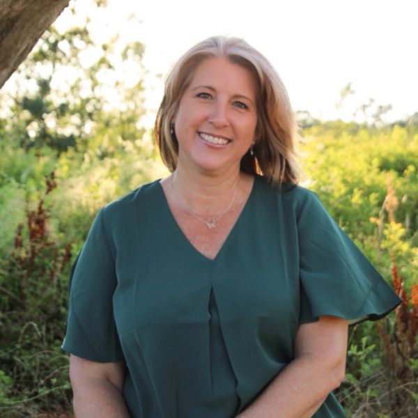 Smiling woman outdoors with greenery in the background.