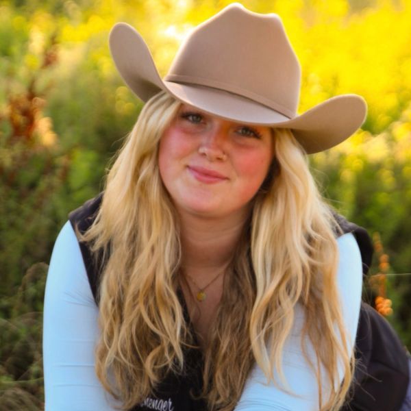 Young woman with long blonde hair wearing a tan cowboy hat outdoors.