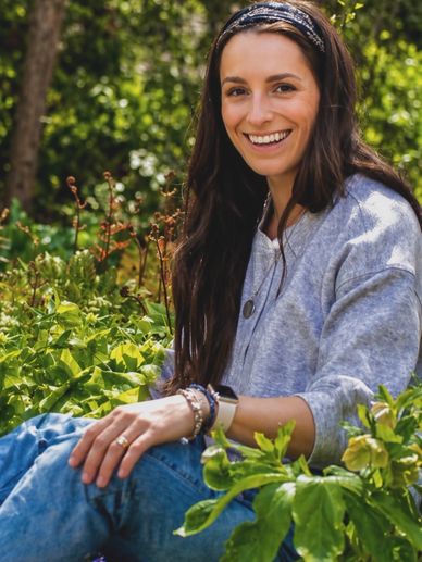Smiling woman sitting outdoors surrounded by greenery.