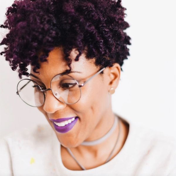 Woman with curly hair and purple lipstick wearing round glasses.