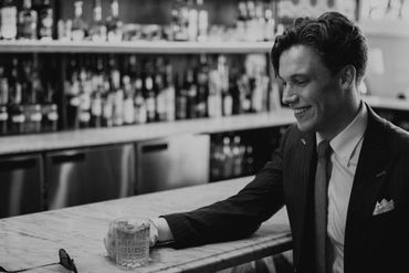 Jared sitting in front of a bar wearing a pinstripe suit and holding a rocks glass.