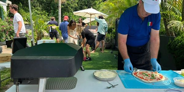 Chef preparing pizza outdoors under a green canopy with guests in the background.