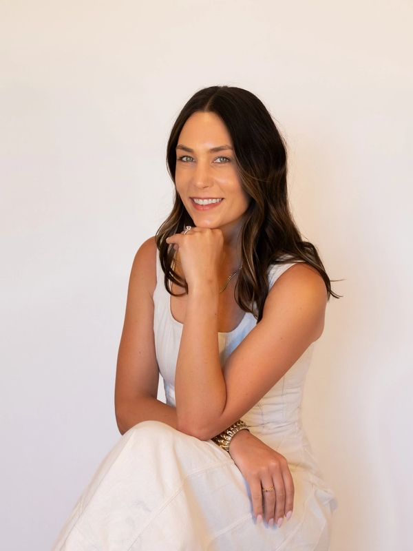 Smiling woman in white dress posing with hand on chin against plain background.