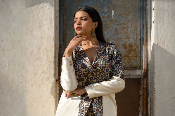 Woman in patterned attire posing thoughtfully against a rustic backdrop.