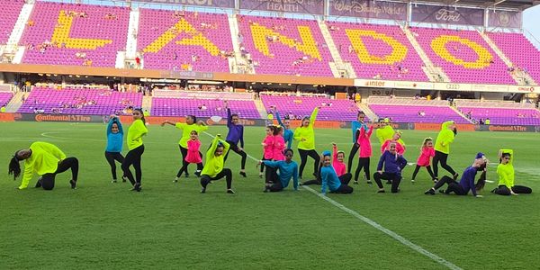 Group of dancers in colorful outfits performing on a soccer field.