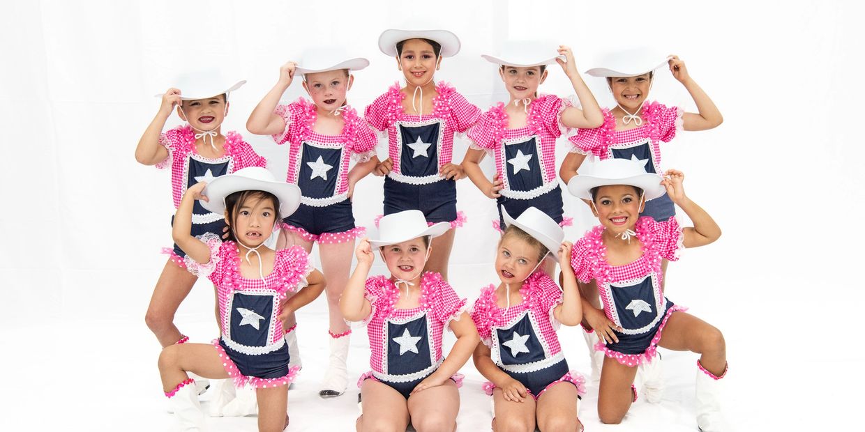 Young girls in pink and navy dance costumes with white cowboy hats pose together.