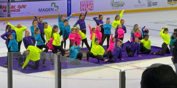 Children in colorful outfits perform a dance routine on ice rink.
