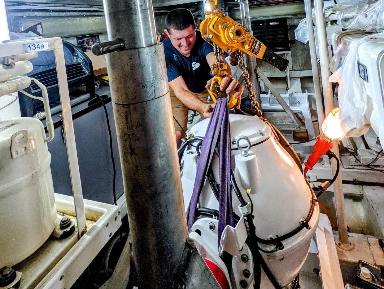 A man operates a hoist to lift a heavy marine engine in a boat's engine room.