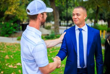 Two men shaking hands outdoors, one in a suit and the other in casual wear.