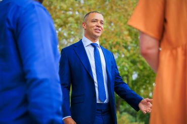 Man in blue suit talking to two people outdoors.