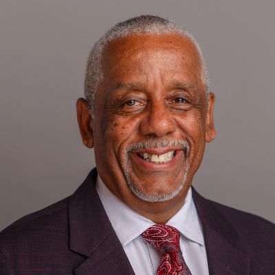 Smiling elderly man in a suit and patterned tie against a plain background.