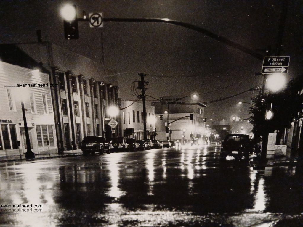 Black and white photo of a wet city street at night with reflections from streetlights.